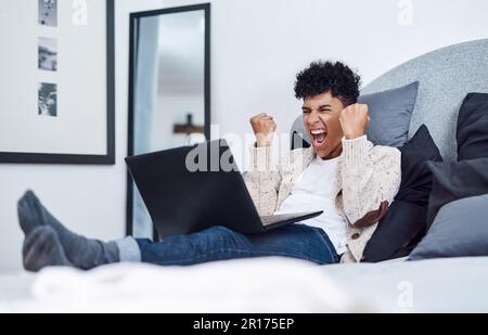 Verrouillé mais pas luqué non. un jeune homme qui applaudit tout en utilisant un ordinateur portable sur son lit à la maison. Banque D'Images