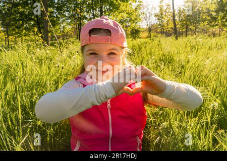 Une jeune fille gaie est assise dans l'herbe verte épaisse, représentant un signe de coeur avec ses mains. Promenez-vous à l'extérieur dans le parc de la ville Banque D'Images