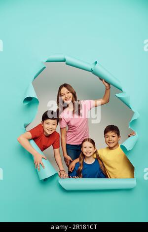 Groupe joyeux d'enfants multiethniques dans des vêtements colorés regardant l'appareil photo pendant la journée de protection des enfants tout en se tenant derrière le trou dans le papier bleu backgrou Banque D'Images