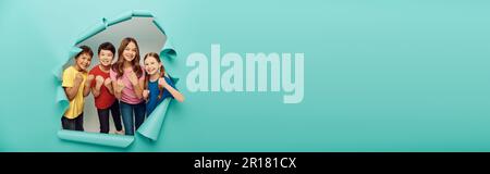 Joyeux gamins multiethniques dans des vêtements colorés montrant des poings pendant la journée internationale d'enfants tout en se tenant derrière le trou dans le fond bleu de papier, Ban Banque D'Images