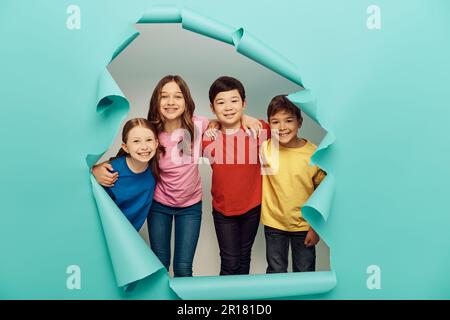 Les enfants souriants de la préadolescence multiethnique dans des t-shirts colorés embrassant pendant la fête de la protection des enfants derrière le trou dans un fond de papier bleu Banque D'Images
