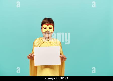 Garçon multiracial souriant en costume de super-héros jaune avec masque tenant du papier blanc sur fond bleu, concept de jour heureux des enfants Banque D'Images