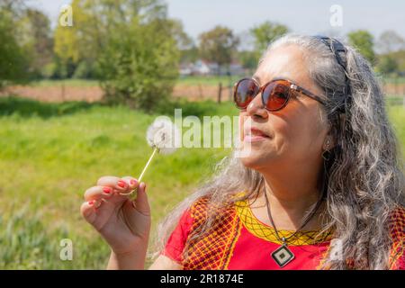 Brunette latino-américaine femme portant des lunettes de soleil regardant la fleur de pissenlit à la main, les arbres dans un fond flou, les cheveux gris bouclés, le bandeau et le nativ Banque D'Images