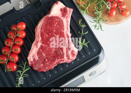 Steaks de première qualité crus sur le gril électrique. Steaks de viande de choix black angus Ribeye sur un grill électrique moderne au romarin, à la tomate cerise et au poivre. Cuisine pro Banque D'Images