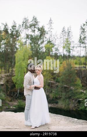 Heureux couple interracial newlyweds se tient sur le rocher et embrasse contre fond de forêt et de canyon. Concept de relations d'amour et d'unité betwee Banque D'Images