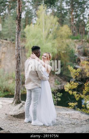 Un couple interracial heureux de jeunes mariés se tient, embrasse et sourit sur fond de lac et de forêt. Concept de relations d'amour et d'unité entre Banque D'Images