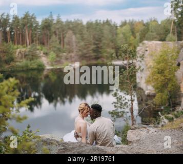 Heureux couple interracial newlyweds se trouve sur la roche et embrasse sur le magnifique fond du lac, de la forêt et du canyon. Concept de relations d'amour an Banque D'Images