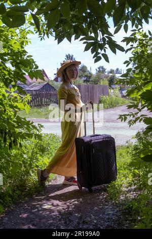 Une fille asiatique dans un chapeau de paille et une robe jaune porte une grande valise noire le long de la route parmi les arbres verts. Ethnotourisme. Le tourisme de village Banque D'Images