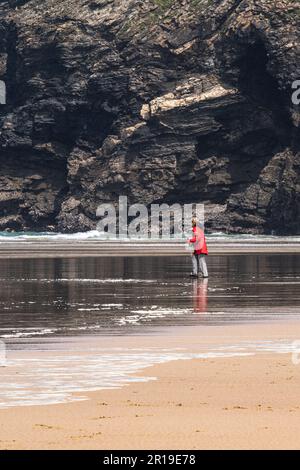 Une femelle mature portant un manteau rouge vif et utilisant des bâtons de marche bâtons de randonnée pédestre marchant sur la rive à la plage de Mawgan Porth dans les Cornouailles au Royaume-Uni. Banque D'Images