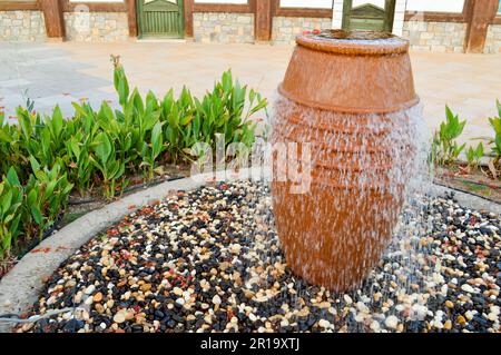 Une belle petite fontaine sous la forme d'un vase brun, une carafe avec des gouttes d'eau tombant sur des pierres de couleur debout dans un lit de fleur avec plante verte Banque D'Images