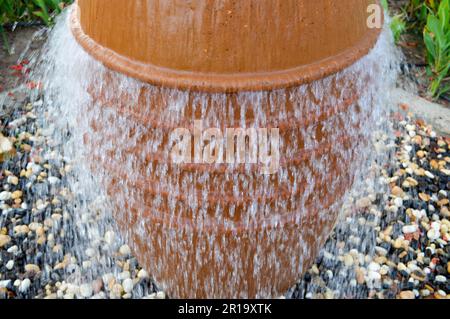 Une belle petite fontaine sous la forme d'un vase brun, une carafe avec des gouttes d'eau tombant sur des pierres de couleur debout dans un lit de fleur avec plante verte Banque D'Images