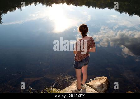 Apocalypse: Jeune femme debout sur un rocher à côté d'un lac avec un reflet lumineux de soleil, New Hampshire, Etats-Unis Banque D'Images