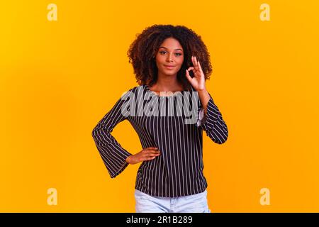 Belle fille à cheveux bouclés en utilisant ses mains ok signe. Arrière-plan jaune. Femme afro ok Banque D'Images