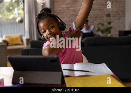 Bonne fille afro-américaine portant des écouteurs levant la main pendant la leçon en ligne à la maison Banque D'Images