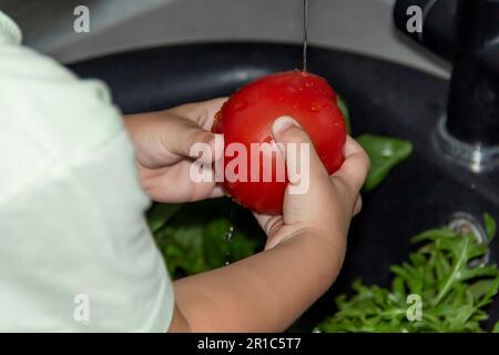 Un petit garçon avec une coupe courte aide à cuisiner dans la cuisine. Lave les légumes frais et les herbes dans un évier noir dans une cuisine grise. Banque D'Images