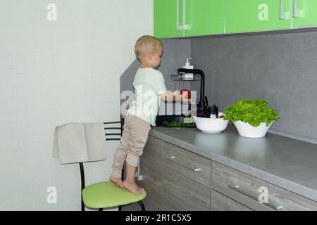 Un petit garçon avec une coupe courte aide à cuisiner dans la cuisine. Lave les légumes frais et les herbes dans un évier noir dans une cuisine grise. Banque D'Images