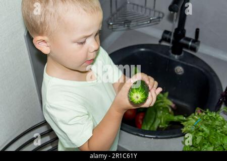 Un petit garçon avec une coupe courte aide à cuisiner dans la cuisine. Lave les légumes frais et les herbes dans un évier noir dans une cuisine grise. Banque D'Images