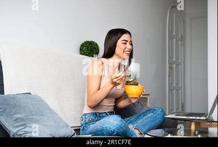 Bonne jeune femme latine mangeant une tasse de céréales avec du lait tout en regardant un film sur un ordinateur portable Banque D'Images