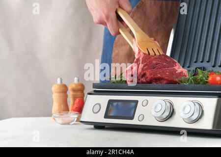 Steaks de première qualité crus sur le gril électrique. Chef à la main tenant le steak Ribeye sur un grill électrique moderne avec romarin, tomates cerises et poivre. Procédure de cuisson Banque D'Images