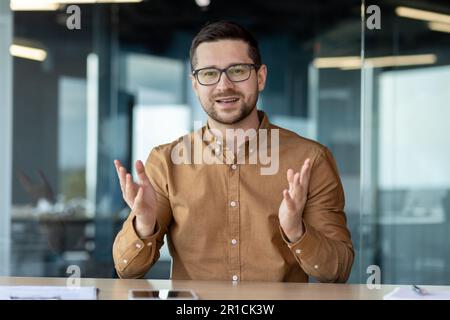 Formation à distance. Jeune homme souriant enseignant, tuteur, formateur assis au bureau, campus devant la caméra et l'enseignement, donnant des conférences, des leçons en ligne par vidéoconférence. Banque D'Images