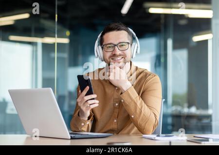 Portrait d'un jeune homme assis dans le bureau au bureau avec un casque, travaillant sur un ordinateur portable et tenant un téléphone. Souriant et regardant avec assurance la caméra, tenant son menton avec sa main. Banque D'Images