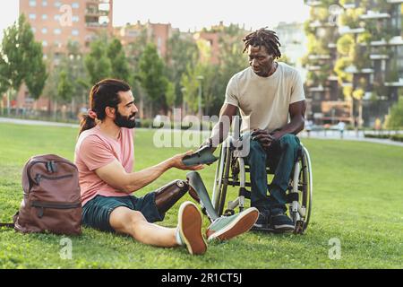 Un hispanique avec une barbe, une queue de cheval et une jambe prothétique se trouve sur l'herbe dans un parc public, montrant un morceau de h à son ami africain en fauteuil roulant Banque D'Images