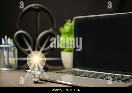 Une ampoule lumineuse et un ordinateur portable sont des écrans noirs vierges sur la table de travail. Concept d'une start-up qui stimule l'économie, la finance, la banque. Casque sans fil Banque D'Images
