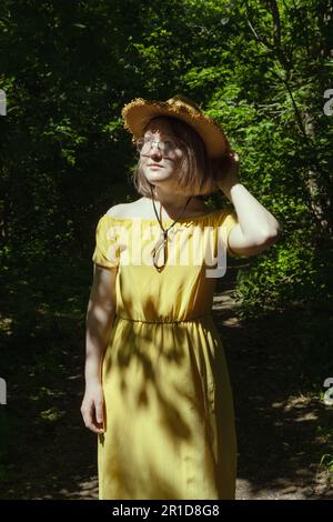 Belle fille asiatique avec verres, chapeau de paille et robe jaune dans la forêt en été, belles ombres, lumière du soleil, portrait. Jeune fille ado rêveuse. Banque D'Images