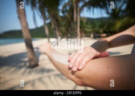 Gros plan de la main pendant l'application de la crème solaire. Soins de la peau pendant les vacances d'été sur une belle plage de sable avec des palmiers dans la destination tropicale. Banque D'Images