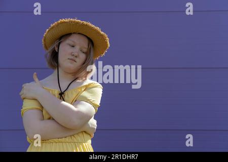 Bouleverser asiatique jeune fille dans un chapeau et la robe se chagre avec ses bras sur un fond violet, la pose fermée, le langage des signes. Banque D'Images