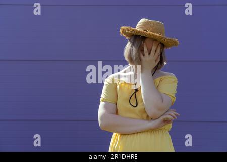 Une jeune fille asiatique frustrée dans un chapeau et une robe couvre son visage avec ses mains sur un fond violet. Concept de santé mentale. Banque D'Images