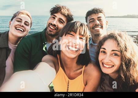 Un groupe diversifié d'amis, y compris une personne non binaire, prennent un selfie sur le front de mer lors d'une chaude journée ensoleillée. Les cinq jeunes adultes, tous dans leur t Banque D'Images