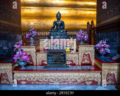 Une statue noire de Bouddha avec des offrandes florales devant une section du Bouddha couché doré au temple Wat Pho à Bangkok, en Thaïlande. Banque D'Images