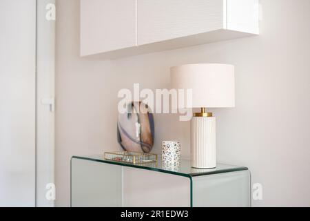 Vue rapprochée d'une console en verre avec accessoires et d'une lampe avec une armoire blanche suspendue dans le couloir ou le salon. Décoration intérieure moderne Banque D'Images