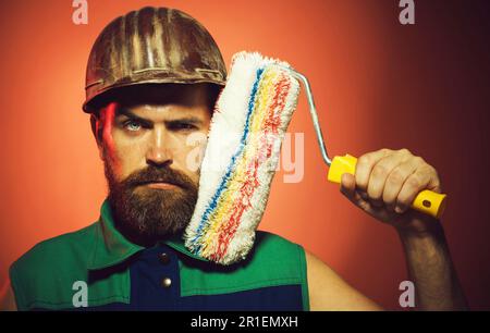 Peintre professionnel homme en casque avec rouleau de peinture. Peintre barbu en uniforme avec outil de peinture. Travail à l'intérieur. Construction et rénovation Banque D'Images