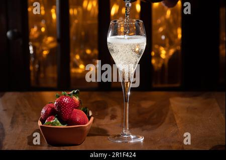 Un verre de champagne à la sauce sautée, des bulles de vin dans un verre de tulipe et un petit bol avec des fraises fraîches sur le bar du soir illuminent le fond Banque D'Images