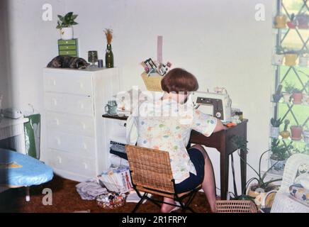 Une femme utilisant une machine à coudre alors qu'un chat dort sur une commode ca. mi-1970s Banque D'Images