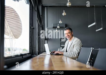 Un homme d'affaires heureux d'un âge moyen assis dans un bureau moderne qui travaille sur un ordinateur portable. Banque D'Images