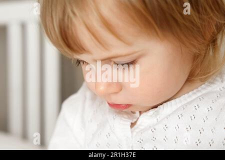 Portrait d'une fille de deux ans avec un regard concentré. Un petit enfant semble mécontent. Blonde 2 ans bébé avec chemise blanche regardant avec le visage triste. Banque D'Images