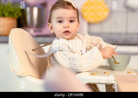 Petite fille assise dans une chaise haute mangeant par elle-même avec une cuillère Banque D'Images