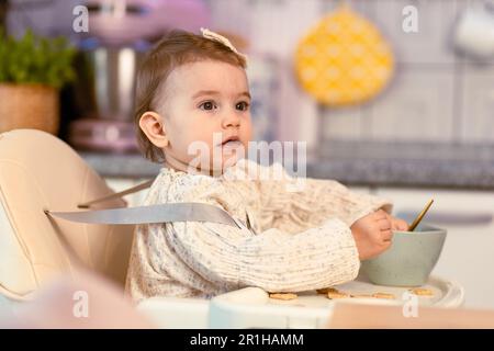 Petite fille assise dans une chaise haute mangeant par elle-même avec une cuillère Banque D'Images