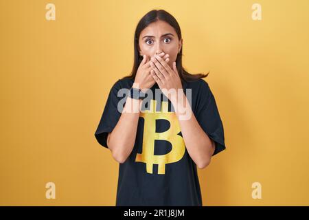 Une jeune femme hispanique portant un t-shirt bitcoin a choqué de couvrir la bouche avec des mains pour erreur. concept secret. Banque D'Images