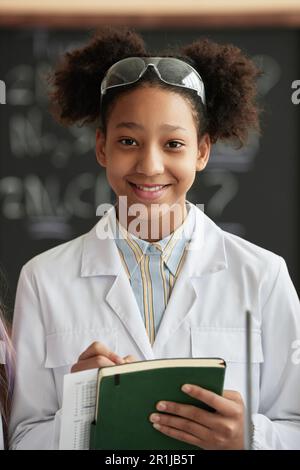 Portrait vertical d'une écolière noire souriante portant un manteau de laboratoire en classe scientifique et regardant un ordinateur portable avec un appareil photo Banque D'Images