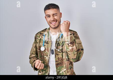 Jeune médecin hispanique portant l'uniforme militaire de camouflage célébrant surpris et étonné de succès avec les bras levés et les yeux fermés Banque D'Images