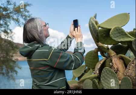 Homme aux cheveux gris avec de longs cheveux photographiant un cactus buisson contre le fond de la mer, Grèce, Crète Banque D'Images