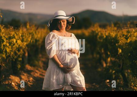 Un portrait d'une femme enceinte dans une robe et un chapeau blancs, tenant doucement son ventre, dans le cadre d'un vignoble, rayonnant la maternité et l'attente Banque D'Images