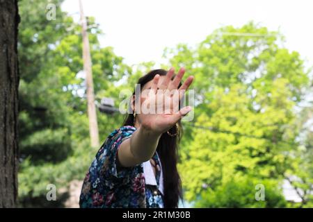 Jeune femme aux cheveux longs et sombres qui tient sa main jusqu'à l'appareil photo, couvrant son visage, décontractée ou peut-être en train de dire arrêt! Robe à imprimé fleuri vintage Banque D'Images