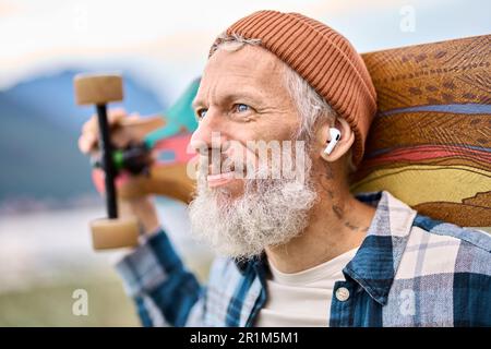 Un homme hippster plus âgé se tenant dans un parc naturel, avec de la musique à l'écoute du skate. Banque D'Images