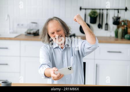 Gai mature caucasien gris cheveux homme avec la barbe, assis à la maison dans la cuisine à la table, reposant, en utilisant son smartphone, joue un jeu vidéo, gagné, gesticulent joyeusement avec sa main, rires Banque D'Images