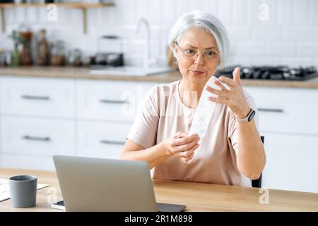 Perplexe, frustrée, cette femme gris caucasien est assise dans la cuisine à une table avec un ordinateur portable, tient une facture entre ses mains, paie des factures en ligne, passe des commandes en ligne, vidée d'une grande quantité Banque D'Images